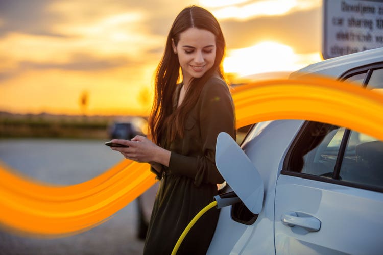 Side view waist up shot of a young woman in a dress using her mobile phone while waiting for her white electric car to charge during sunset